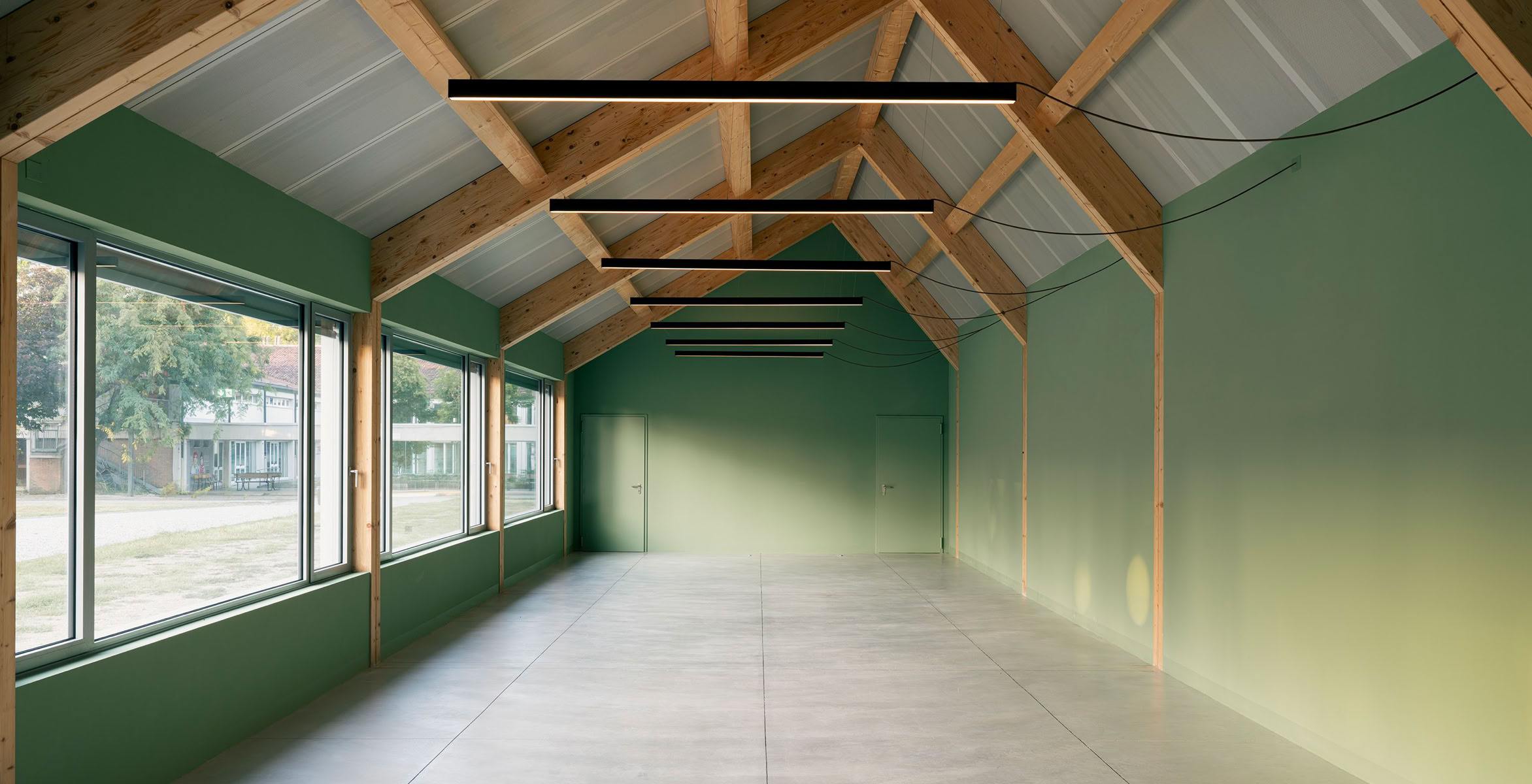 Interno della mensa scolastica di Villa del Conte con pareti verdi, soffitto con travi in legno e grandi finestre sul lato sinistro.